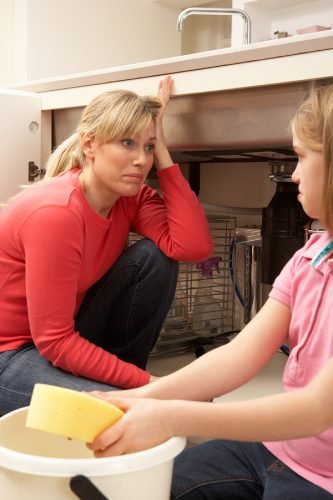 Woman crouches down under leaking kitchen sink with young daughter nearby helping