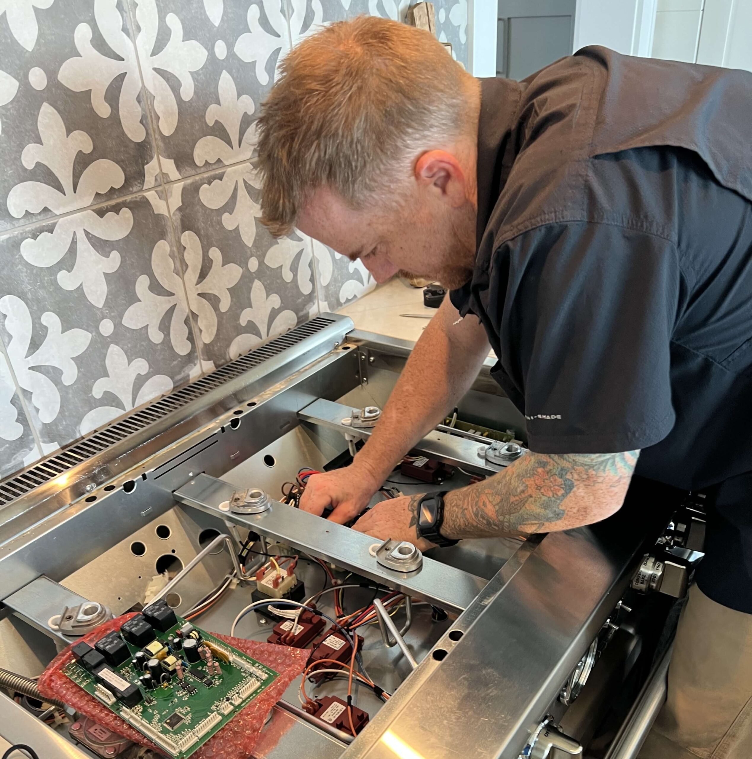 A man wearing a black shirt is inspecting or repairing a stove in a narrow kitchen corridor.