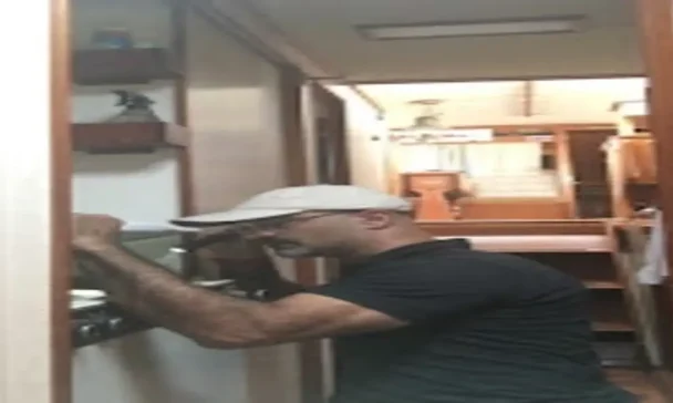 A man wearing a white cap and black shirt is inspecting or repairing a stove in a narrow kitchen corridor. The background shows wooden cabinets and shelves, creating a compact kitchen environment.