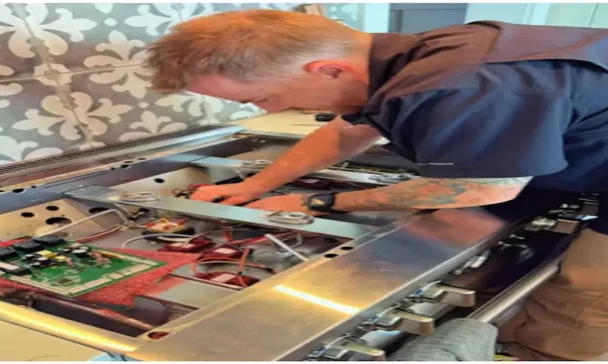 A technician with a tattooed arm is repairing the internal components of a stove. The cooktop is lifted open, revealing circuit boards and wiring, indicating an in-depth stove maintenance task.