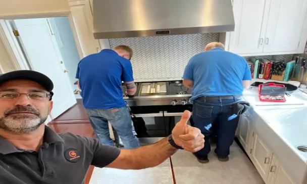 A man in a cap and glasses gives a thumbs-up while two technicians in blue shirts work on a stove in a modern kitchen. Tools and books are visible on the countertop.